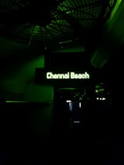 A dimly lit sign displays the words 'Chennai Beach' in glowing green text. The background is largely dark, with some faint outlines of structural elements and shadows visible.