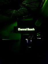 A dimly lit sign displays the words 'Chennai Beach' in glowing green text. The background is largely dark, with some faint outlines of structural elements and shadows visible.
