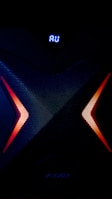 A dark, high-tech speaker with a futuristic design, featuring a prominent X pattern with red and orange lighting accents. Above the X pattern, a digital display shows the letters 'AU' in bright blue.