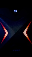 A dark, high-tech speaker with a futuristic design, featuring a prominent X pattern with red and orange lighting accents. Above the X pattern, a digital display shows the letters 'AU' in bright blue.