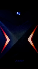 A dark, high-tech speaker with a futuristic design, featuring a prominent X pattern with red and orange lighting accents. Above the X pattern, a digital display shows the letters 'AU' in bright blue.