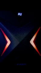 A dark, high-tech speaker with a futuristic design, featuring a prominent X pattern with red and orange lighting accents. Above the X pattern, a digital display shows the letters 'AU' in bright blue.