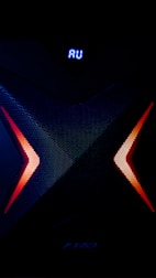 A dark, high-tech speaker with a futuristic design, featuring a prominent X pattern with red and orange lighting accents. Above the X pattern, a digital display shows the letters 'AU' in bright blue.
