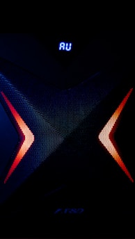 A dark, high-tech speaker with a futuristic design, featuring a prominent X pattern with red and orange lighting accents. Above the X pattern, a digital display shows the letters 'AU' in bright blue.