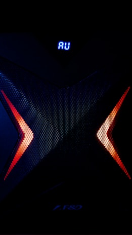 A dark, high-tech speaker with a futuristic design, featuring a prominent X pattern with red and orange lighting accents. Above the X pattern, a digital display shows the letters 'AU' in bright blue.