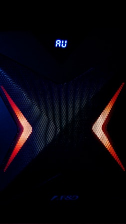 A dark, high-tech speaker with a futuristic design, featuring a prominent X pattern with red and orange lighting accents. Above the X pattern, a digital display shows the letters 'AU' in bright blue.