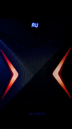 A dark, high-tech speaker with a futuristic design, featuring a prominent X pattern with red and orange lighting accents. Above the X pattern, a digital display shows the letters 'AU' in bright blue.