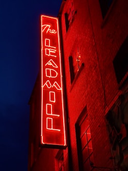 A vibrant red neon sign reading 'The Leadmill' is mounted on the brick exterior of a building. The sign glows against the dark blue night sky, creating a striking contrast. The building features several windows with darkened glass.