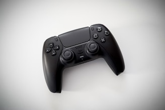 A black gaming controller with dual thumbsticks and multiple buttons sits on a light background. The design is modern and ergonomic, featuring a glossy finish and familiar button layout with directional and action buttons.