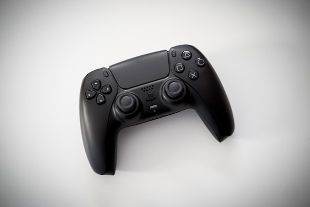 A black gaming controller with dual thumbsticks and multiple buttons sits on a light background. The design is modern and ergonomic, featuring a glossy finish and familiar button layout with directional and action buttons.