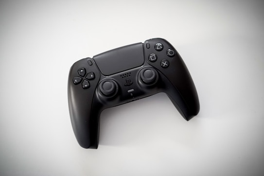 A black gaming controller with dual thumbsticks and multiple buttons sits on a light background. The design is modern and ergonomic, featuring a glossy finish and familiar button layout with directional and action buttons.