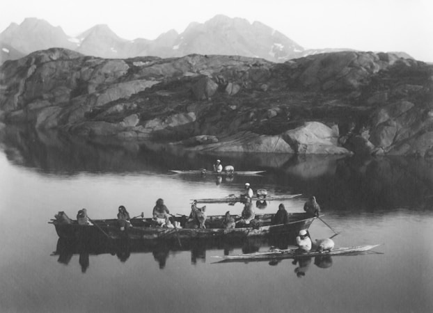 A group of individuals are paddling traditional boats in a calm, reflective body of water. The backdrop features rugged rocky terrain and distant mountains, creating a serene and natural setting.