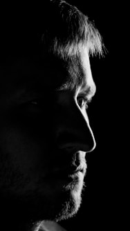 A close-up black and white portrait of a person's face in profile. The image highlights strong contrasts with dramatic lighting, focusing on the eye and facial features, while most of the background and side of the face are in shadow.