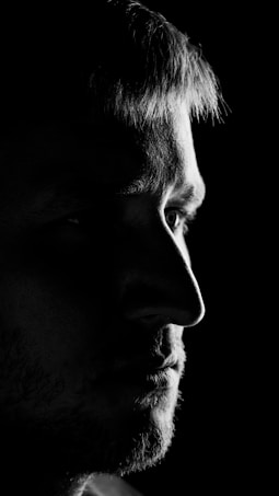 A close-up black and white portrait of a person's face in profile. The image highlights strong contrasts with dramatic lighting, focusing on the eye and facial features, while most of the background and side of the face are in shadow.