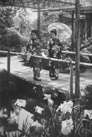 Two women dressed in traditional kimonos stand on a wooden bridge in a serene Japanese garden. One of the women holds a parasol. The garden is lush with trees and flowers, and there is a small body of water beneath the bridge with blooming irises in the foreground.