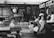 A historical classroom setting with several students seated at wooden desks. The teacher stands at the front near a blackboard filled with writings and diagrams. A globe is placed on a table, and educational charts hang on the walls.