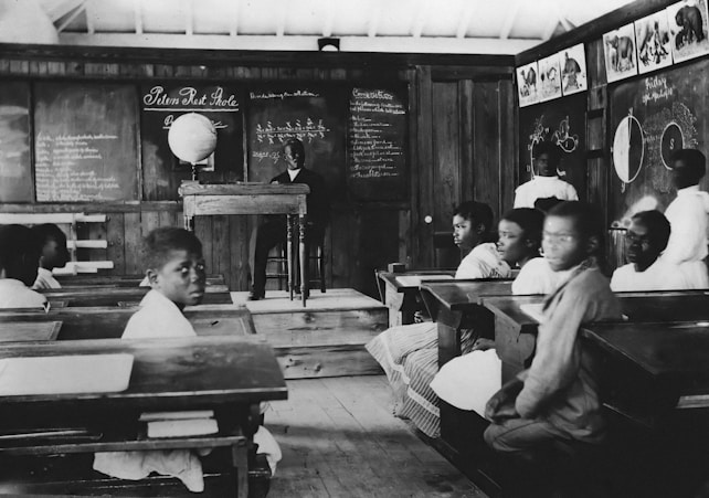 A historical classroom setting with several students seated at wooden desks. The teacher stands at the front near a blackboard filled with writings and diagrams. A globe is placed on a table, and educational charts hang on the walls.