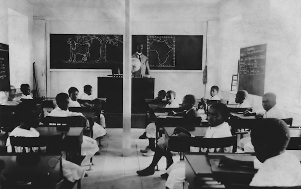 A classroom setting with several students seated at individual desks, all facing a teacher standing near a blackboard. The room has simple furniture and chalkboards with illustrations, including a map of Africa and drawings of a cow and tree.