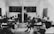 A classroom setting with several students seated at individual desks, all facing a teacher standing near a blackboard. The room has simple furniture and chalkboards with illustrations, including a map of Africa and drawings of a cow and tree.