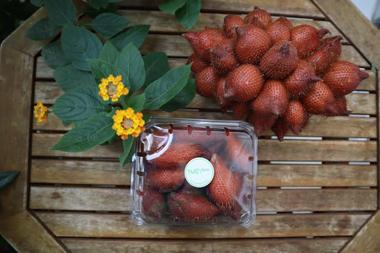 A collection of reddish-brown snake fruits is placed on a wooden surface alongside a plastic container filled with more of the same fruits. Bright yellow flowers and green leaves are situated next to the fruits, adding a splash of color to the scene.