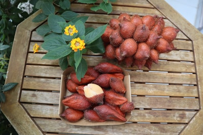 A wooden table holds a cluster of red-brown, scaly fruits, with one fruit partially peeled to reveal its pale interior. Next to it, a leafy green plant with small yellow flowers adds a contrasting burst of color.
