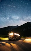 A van is parked on a road by the countryside at night, with bicycles mounted on the back. The vehicle is illuminated from within, casting a warm glow on the surrounding area. Above, a clear starry sky is visible, with a shooting star or satellite streaking across.