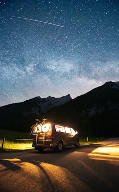 A van is parked on a road by the countryside at night, with bicycles mounted on the back. The vehicle is illuminated from within, casting a warm glow on the surrounding area. Above, a clear starry sky is visible, with a shooting star or satellite streaking across.