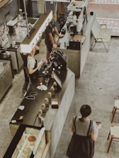 Inside a modern café, there are people interacting at a counter. Various items, including cups, a cash register, and a coffee machine, occupy the counter. The setup is industrial with concrete surfaces and minimalistic design. A large sign reading 'CASHIER' is prominently visible above the counter.
