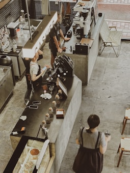 Inside a modern café, there are people interacting at a counter. Various items, including cups, a cash register, and a coffee machine, occupy the counter. The setup is industrial with concrete surfaces and minimalistic design. A large sign reading 'CASHIER' is prominently visible above the counter.