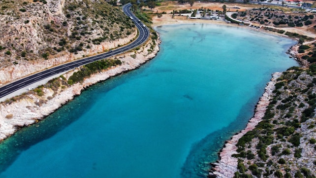 A winding coastal road runs along a turquoise bay with clear waters. The rocky terrain is dotted with sparse vegetation, and several vehicles are visible on the road. In the background, a beach area with structures and trees can be seen, adding to the serene coastal landscape.