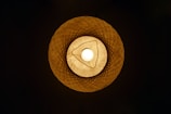 A warm-colored, woven lampshade with a bright light bulb in the center, creating an intricate and symmetrical pattern of shadows and light.