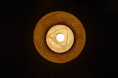 A warm-colored, woven lampshade with a bright light bulb in the center, creating an intricate and symmetrical pattern of shadows and light.