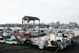 Numerous small boats are docked at a pier with a cityscape in the background. The boats are filled with various items and equipment, and a pavilion structure stands prominently among them. The city across the water features diverse buildings, including residential and commercial structures.