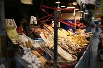 A bustling night market stall displaying a variety of street food. The food items are arranged in large round trays and baskets atop a metal counter illuminated by hanging lights. Skewered meats, fried snacks, and possibly seafood are visible. The stall is adorned with handwritten signs containing Chinese characters. A vendor wearing a face mask stands to the left, while a customer in the background is looking at their phone.