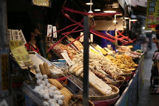 A bustling night market stall displaying a variety of street food. The food items are arranged in large round trays and baskets atop a metal counter illuminated by hanging lights. Skewered meats, fried snacks, and possibly seafood are visible. The stall is adorned with handwritten signs containing Chinese characters. A vendor wearing a face mask stands to the left, while a customer in the background is looking at their phone.