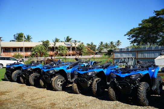 A row of blue all-terrain vehicles (ATVs) is parked on a gravel surface. In the background, there is a building with a beige roof and wooden railings, surrounded by tropical palm trees and other greenery. The sky is clear and blue, creating a bright and sunny atmosphere.