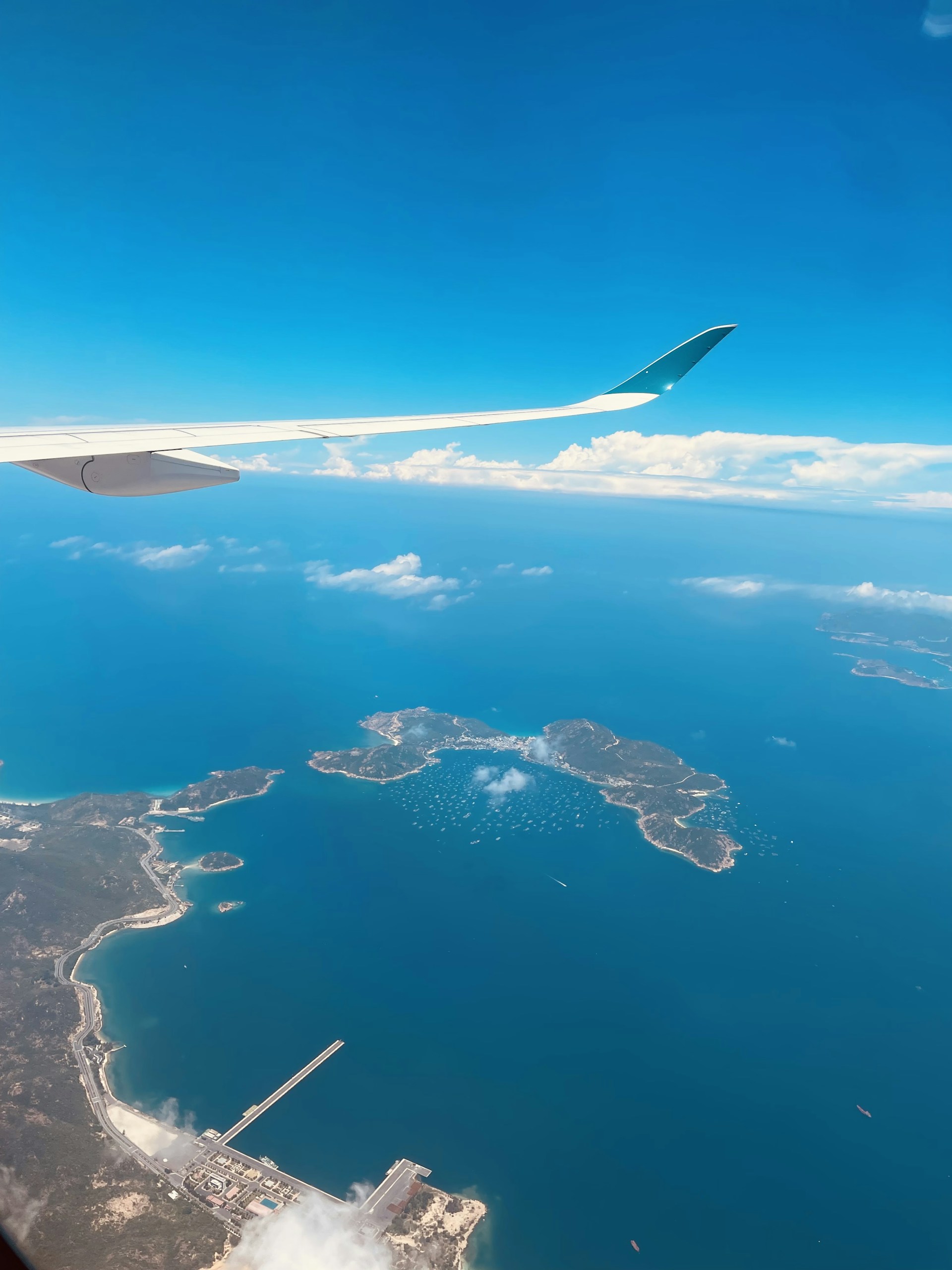 An aerial view displays a vast expanse of blue ocean with islands dotting the water. A section of an airplane wing is visible in the top left, suggesting the photo was taken mid-flight. Cloud formations are scattered above and around the islands, with a clear distinction between the deep blue sea and the sky.