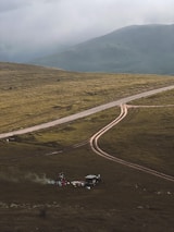 A rugged, grassy landscape with a dirt road curving through it. In the foreground, a small campsite is set up with a vehicle nearby and some camping gear scattered around. Smoke rises from a small fire, creating a serene and remote atmosphere. Mountains loom in the background under a cloudy sky.
