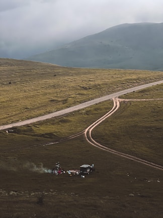 A rugged, grassy landscape with a dirt road curving through it. In the foreground, a small campsite is set up with a vehicle nearby and some camping gear scattered around. Smoke rises from a small fire, creating a serene and remote atmosphere. Mountains loom in the background under a cloudy sky.
