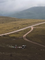 A rugged, grassy landscape with a dirt road curving through it. In the foreground, a small campsite is set up with a vehicle nearby and some camping gear scattered around. Smoke rises from a small fire, creating a serene and remote atmosphere. Mountains loom in the background under a cloudy sky.