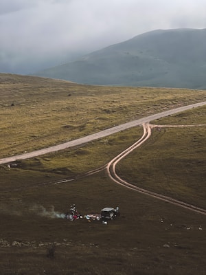 A rugged, grassy landscape with a dirt road curving through it. In the foreground, a small campsite is set up with a vehicle nearby and some camping gear scattered around. Smoke rises from a small fire, creating a serene and remote atmosphere. Mountains loom in the background under a cloudy sky.