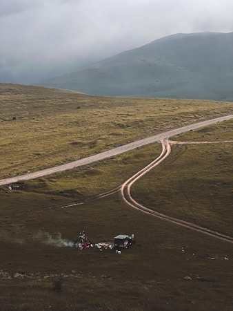 A rugged, grassy landscape with a dirt road curving through it. In the foreground, a small campsite is set up with a vehicle nearby and some camping gear scattered around. Smoke rises from a small fire, creating a serene and remote atmosphere. Mountains loom in the background under a cloudy sky.
