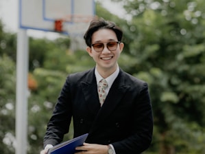 A person wearing sunglasses and a black suit, holding a folder or book, is smiling in front of a background with blurred greenery and a basketball hoop.