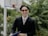 A person wearing sunglasses and a black suit, holding a folder or book, is smiling in front of a background with blurred greenery and a basketball hoop.