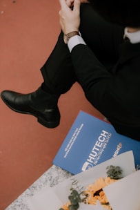A person in formal attire with a wristwatch is seated, with focus on their hands and legs. Nearby, a blue folder labeled 'HUTECH' is placed on a surface, along with a bouquet of yellow flowers.