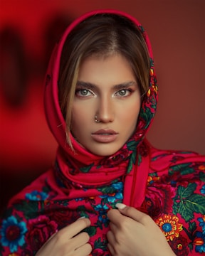 A person gazes directly at the camera, wearing a vibrant, floral-patterned scarf in shades of red, blue, and green. The scarf covers their head, creating a striking contrast with their bright eyes and neutral makeup tones. The background is softly blurred with a warm red hue, enhancing the focus on the subject.