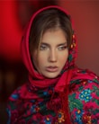 A person is wearing a vibrant, floral-patterned scarf with a red base and colorful flowers in blue, green, and yellow. The scarf is draped around the head, accentuating the person's features. The background is a warm shade of red, enhancing the vivid colors of the scarf.