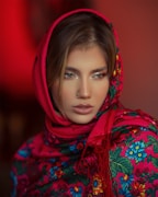 A person is wearing a vibrant, floral-patterned scarf with a red base and colorful flowers in blue, green, and yellow. The scarf is draped around the head, accentuating the person's features. The background is a warm shade of red, enhancing the vivid colors of the scarf.