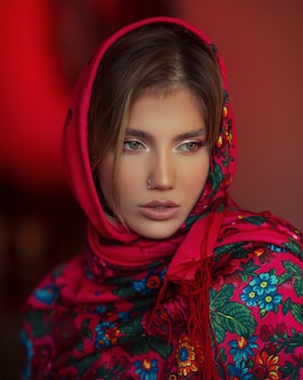 A person is wearing a vibrant, floral-patterned scarf with a red base and colorful flowers in blue, green, and yellow. The scarf is draped around the head, accentuating the person's features. The background is a warm shade of red, enhancing the vivid colors of the scarf.