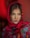 A person is wearing a vibrant, floral-patterned scarf with a red base and colorful flowers in blue, green, and yellow. The scarf is draped around the head, accentuating the person's features. The background is a warm shade of red, enhancing the vivid colors of the scarf.
